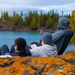 Three interns wearing hoodies and hats sit facing away from the camera, leaning against a rock covered in orange lichen, starkly contrasted against the blue water of Lake Superior and bright evergreen trees in the background.