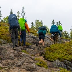 Five students with backpacks, wearing hiking gear, trek up a steep rocky hillside dotted with grass and bushes on a cloudy day.