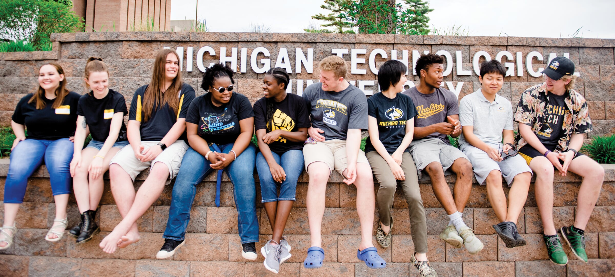 Nine smiling people sit on a brick ledge in front of a sign reading Michigan Technological University.