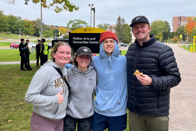 Four people stand close together outside while one eats a piece of pie.