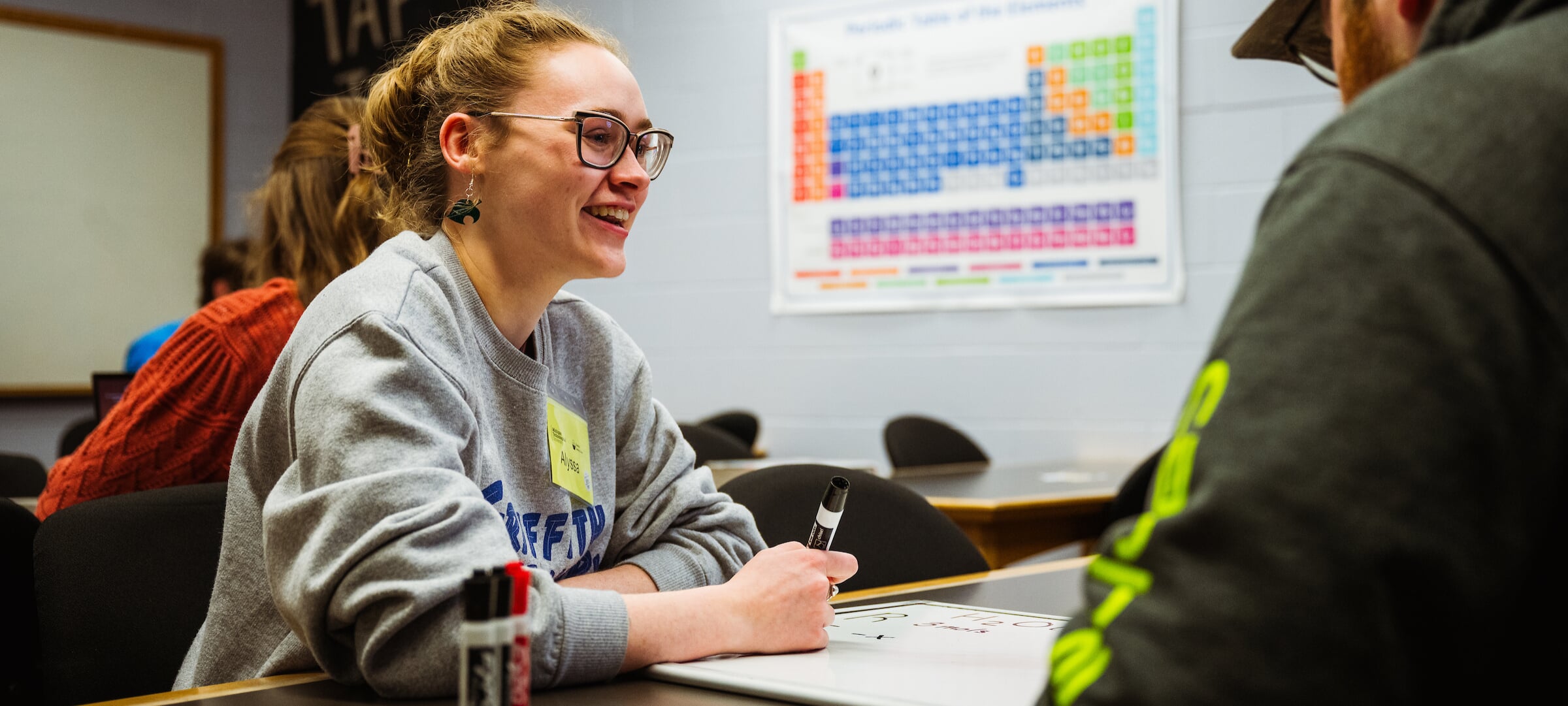 A learning center coach helps a student while both sit at a table in front of a periodic table of elements.