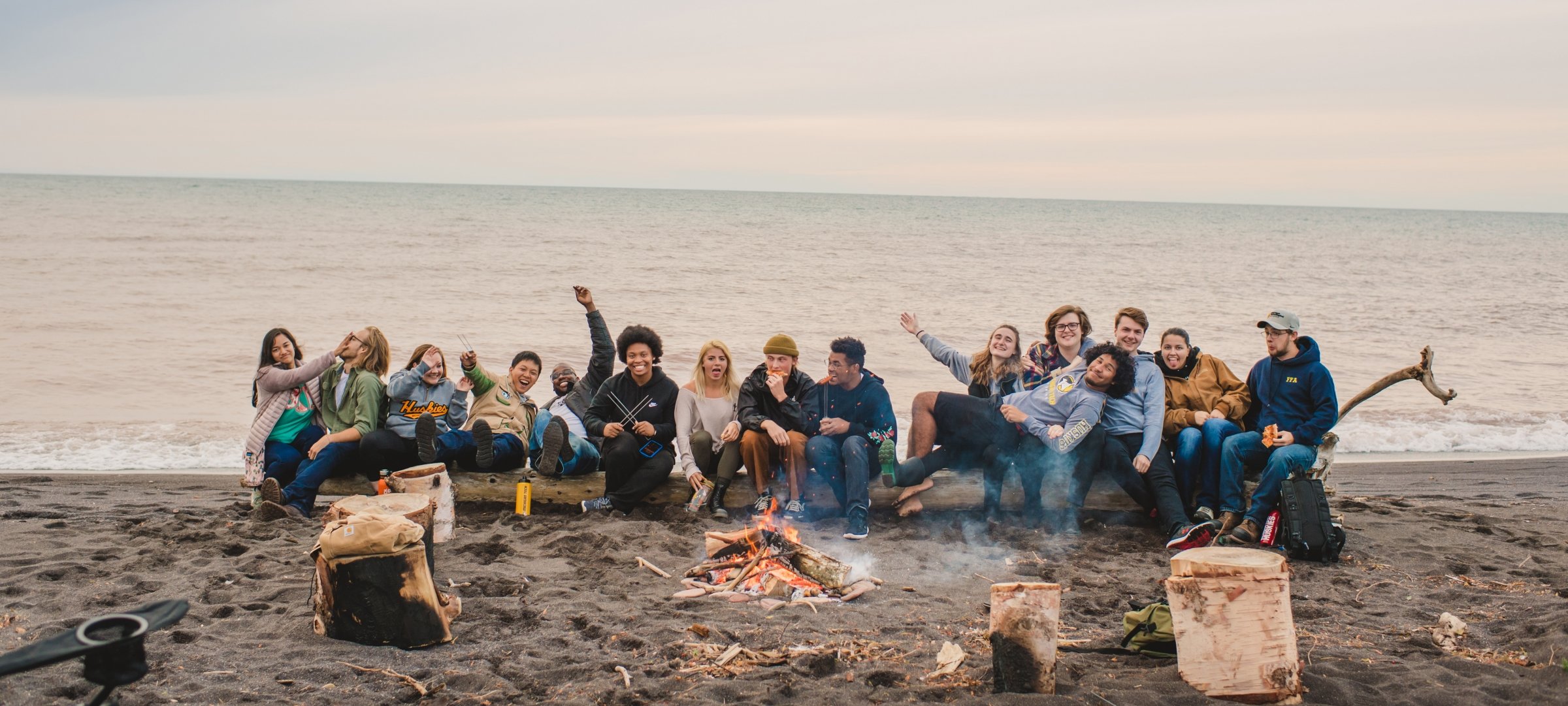 A diverse group of laughing students sits together on a log on a sandy beach by a campfire.