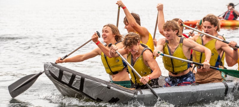 Students wearing life jackets paddling a cardboard boat.