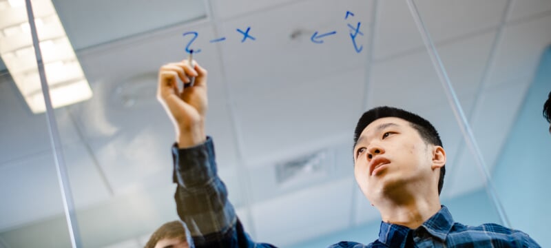 Professor writing on a clear board with a dry-erase marker.