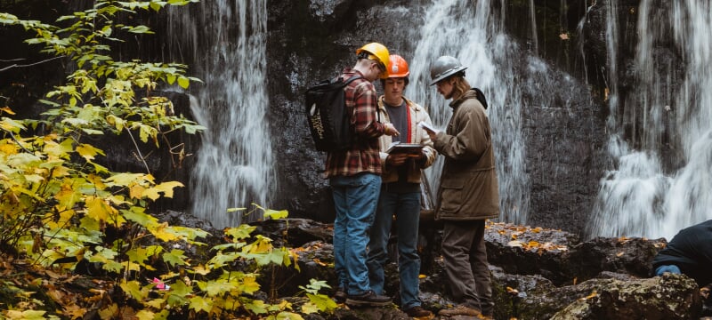 Three people in hard hats standing on rocks in front of a waterfall.