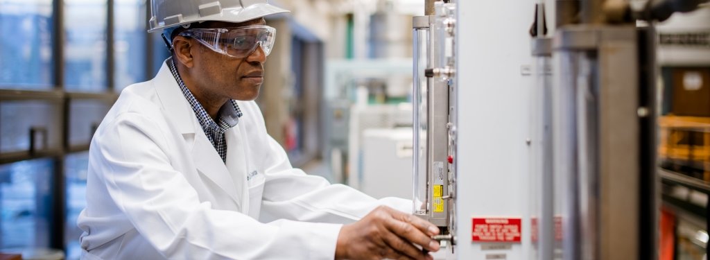 A man, Andre Da Costa, wearing safety googles, a hard hat, and a white lab coat turns a knob on a machine in a research lab. 