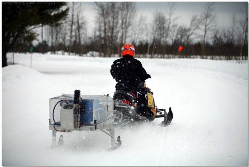 A snowmobile tows a mobile acoustics and emissions testing device mounted on two ski-like runners.