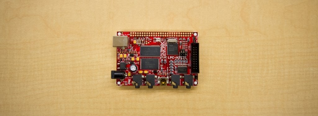 A red circuit board sits on a blond wood table. 