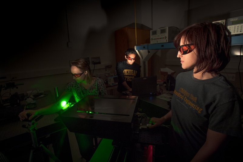 Three people stand around a box-like piece of equipment lit only by the green light of a laser