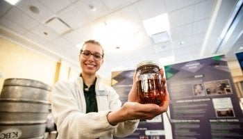 a young woman holding a glass of amber liquid in front of a poster in a ballroom exhibit with a silver keg in the background she is wearing glasses