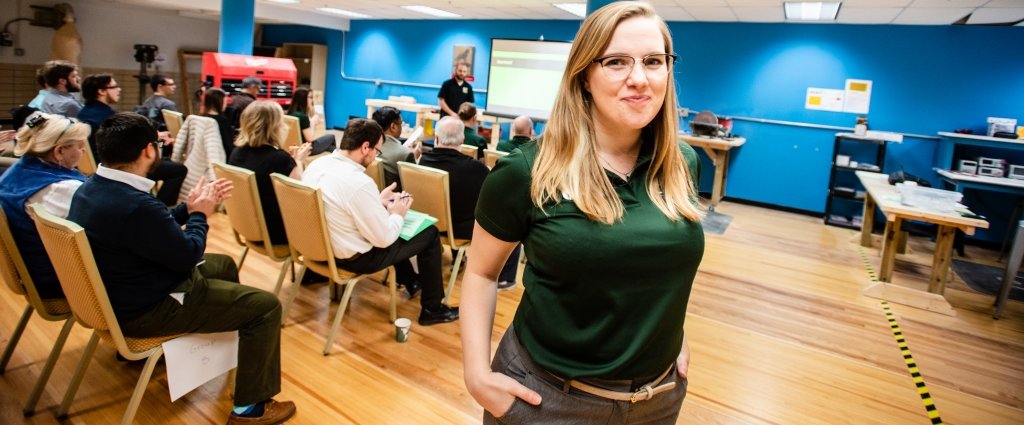 A young woman in a green shirt stands in the foreground of a presentation with the seated audience and a young man presenting in front of a multimedia screen in a makerspace behind her