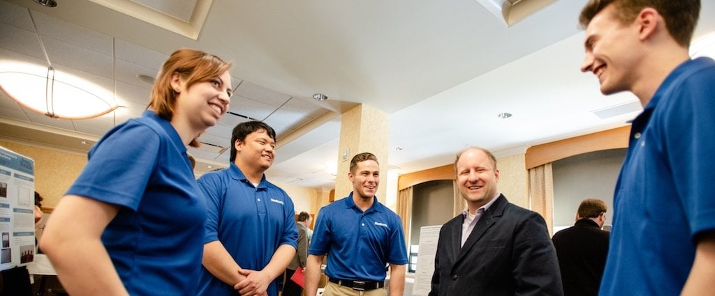 A young woman and three young men all in blue shirts talk with a businessman in a suit in a ballroom with a poster in the background