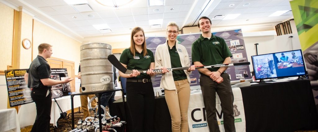Three young people two women and a man hold a hockey stick with a video screen in the background and a beer keg and another young man in the background near a poster 