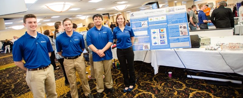 Four people with blue shirts on standing in front of a table in a ballroom with posters and a display