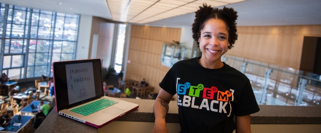 a young woman with a computer in a library on a floor overlooking another floor where students are studying at tables. 