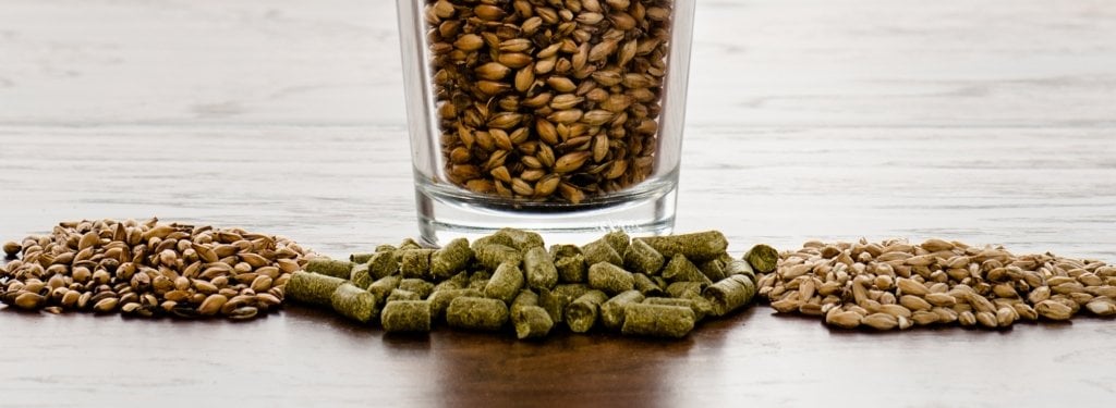 A pint glass filled with malt and piles of mile and pelletized hops in front of the glass on a counter top.