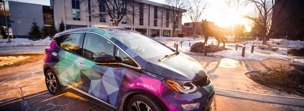 A car with a bright wrap of aurora borealis colors is backlit by the setting sun on Michigan Tech's campus