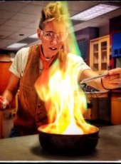 A woman in safety glasses leans over a lab bench with a beaker spouting green and orange flames.
