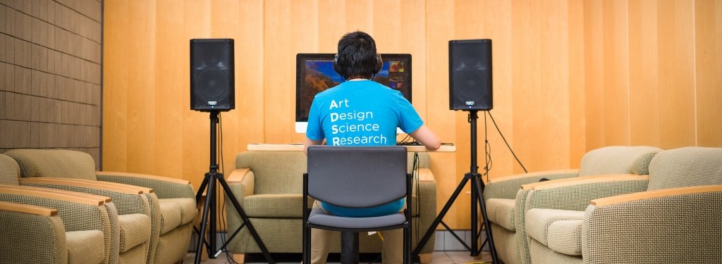 person sitting at a computer with large speakers on either side wearing a shirt that says Art Design Science Research