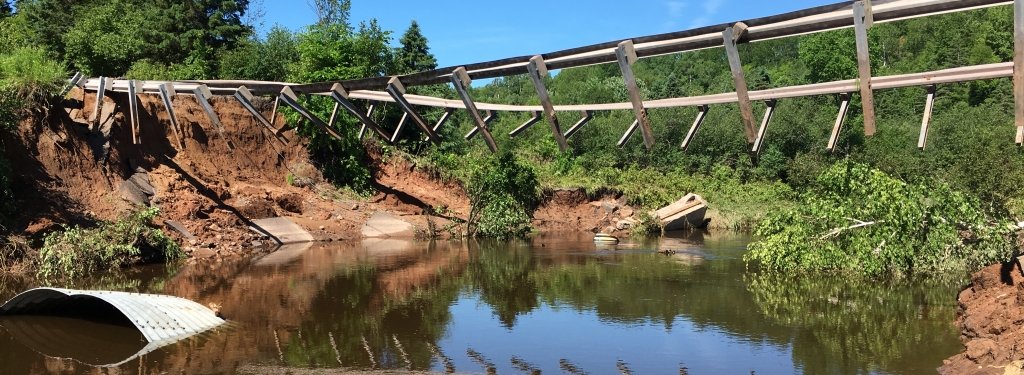 Washed-out bridge supports hang suspended over a river. A washed-out culvert floats in the water.