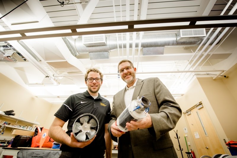 two men hold pieces of equipment including a clear tube and a plate-sized metal fan