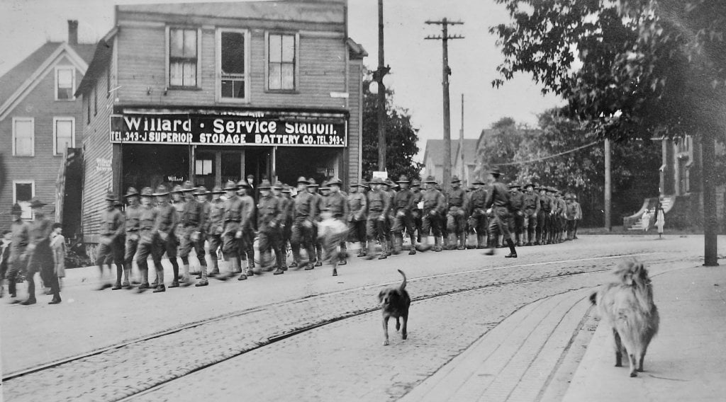 200 World War I era soldiers march down a cobbled street in 1918 with two dogs in the foreground, one on a sidewalk, and a sign on a storefront in the background that says Willard Service Station.  