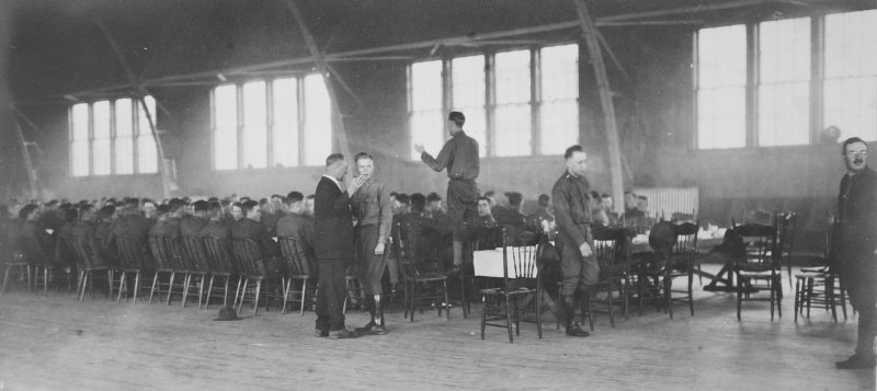 a group of men in an armory sitting at tables eating with one man directing singing from 1918 black and white