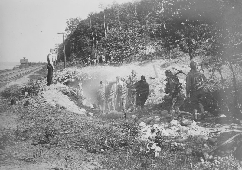 A man in a hat and shirtsleeves overseeing men digging into a rocky hillside with shovels, on the side of a canal, in 1918.
