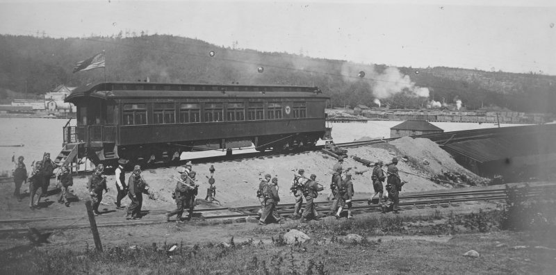 A train car on the side of a hill next to a canal outside with soldiers in training wearing gas masks from 1918 black and white