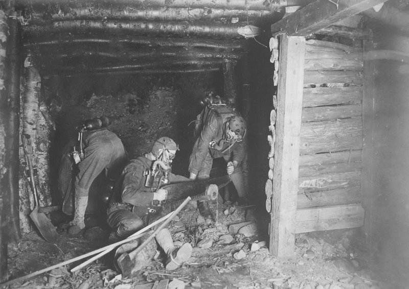 Three men in 1918 with gas masks on in a mine with timbers over their heads holding a vintage drill