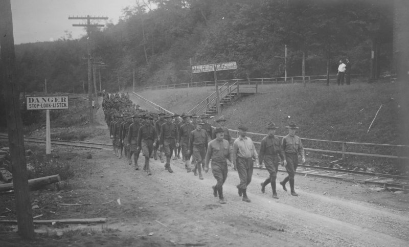men in soldier uniforms march along a black sand path with a stop look and listen danger side outside in formation