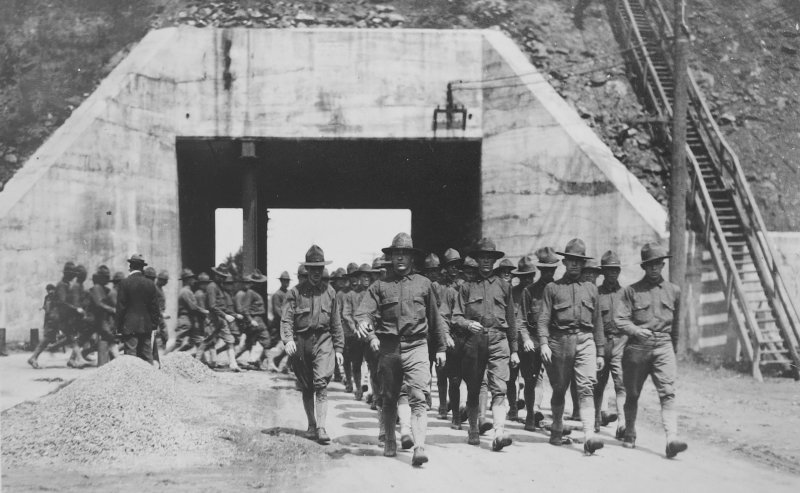 Soldiers in uniform march with an arch behind them at a graveled mine site outdoors in 1918