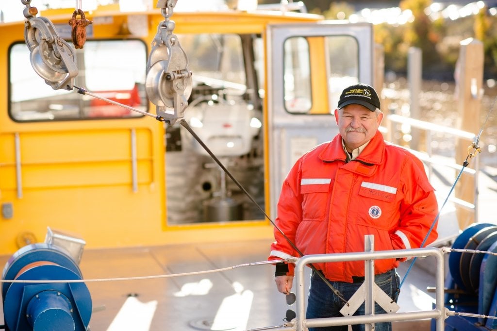 A man in an orange life jacket on a boat at a dock with a steel floor and pilothouse in the background and pulleys in the foreground