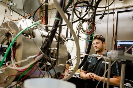 Cords, tubes and wires come out from a chrome cylinder shown in the foreground. A man wearing safety goggles observes in the background.