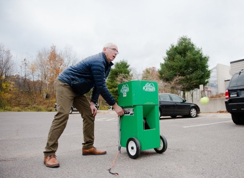 A man triggers the handle on a tennis ball cannon outdoors in a parking lot in the fall in Michigan. The tennis ball shoots through the air. There are cars parked in the background.