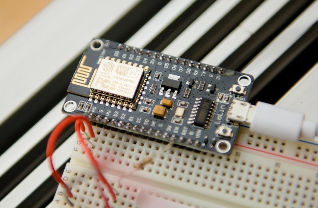 A circuit board connected to a larger circuit board by red wires, sitting on a steel-colored radiator vent. It is a device prototype to detect and ultimately prevent bird-window collisions