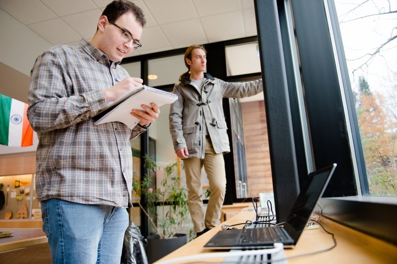 Two young men stand at a window overlooking a parking lot, with the man in the foreground recording data from a laptop in a spiral-bound notebook and the student in the background looking outside.