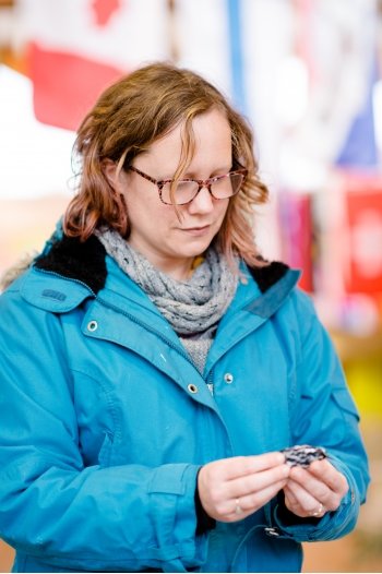 A woman in a blue jacket, wearing glasses, gazes down at a small black and white bird she is holding in her hands. It's dead. She is inside a building on a college campus as part of an experiment being conducted to curb bird-window collisions.