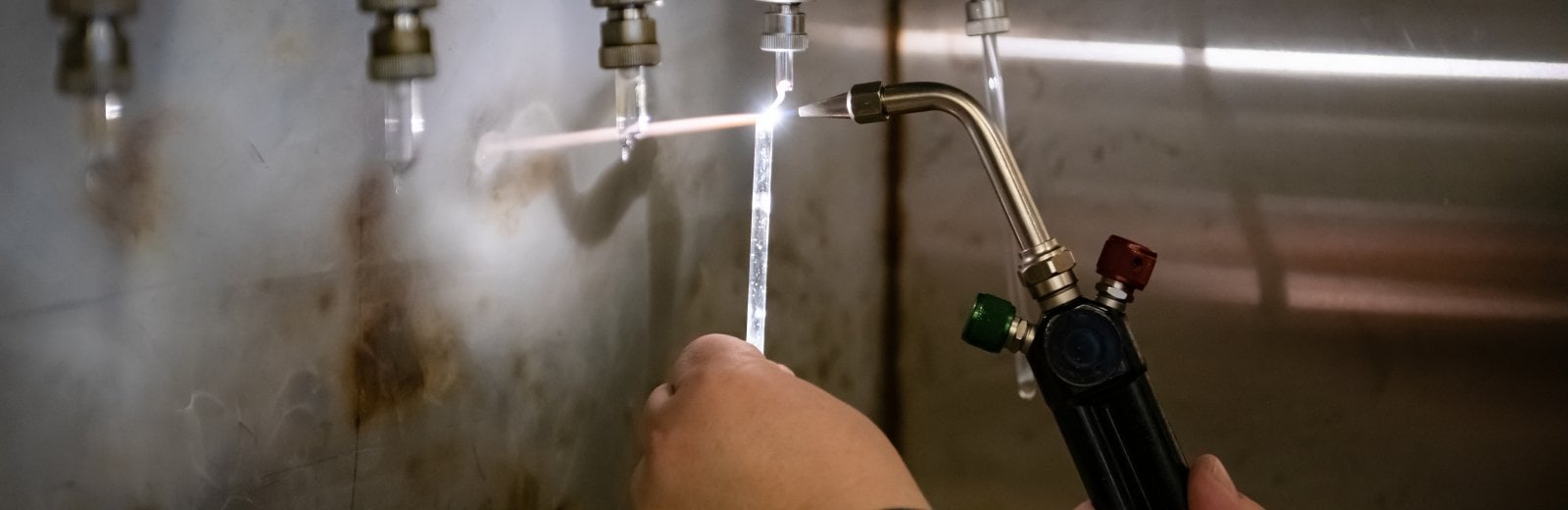 A pair of hands holding a hydrogen-oxygen torch, which is emitting a bright white flame against a slender glass vial.