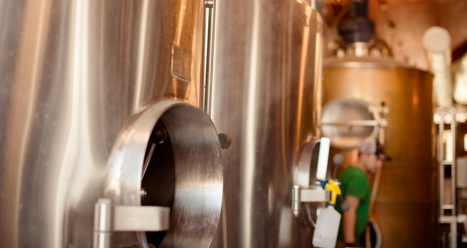 Fermentation vats at an indoor brewery with a brewer in a green shirt in the background. 