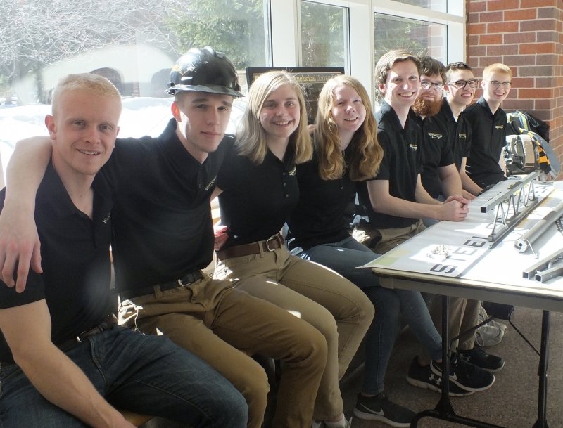 A row of young men and women in Michigan Tech Steel Bridge polo shirts and khaki pants with bests sitting in front of a sunlit window with snow visible outside and a table in front of them with info about the team.