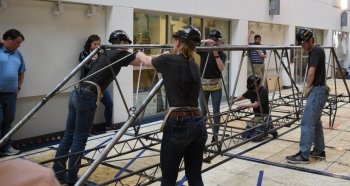 students in black build a steel bridge consisting of pipes and bolts with mirrors behind them and older adults watching in a gym. 