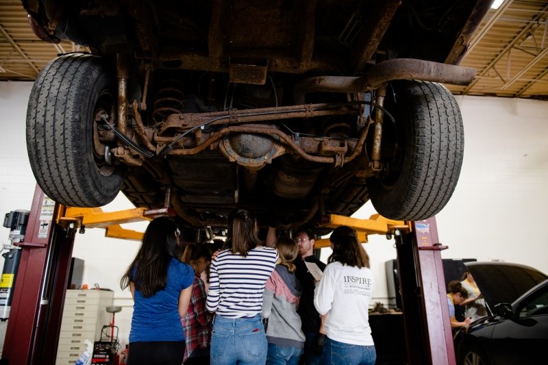 group of young women standing under a vehicle lifted on a hydraulic lift