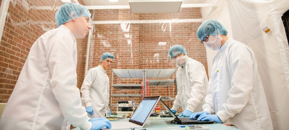 four capped and gloved white-coated students gather in a plastic-draped clean room with brick walls in a University lab.