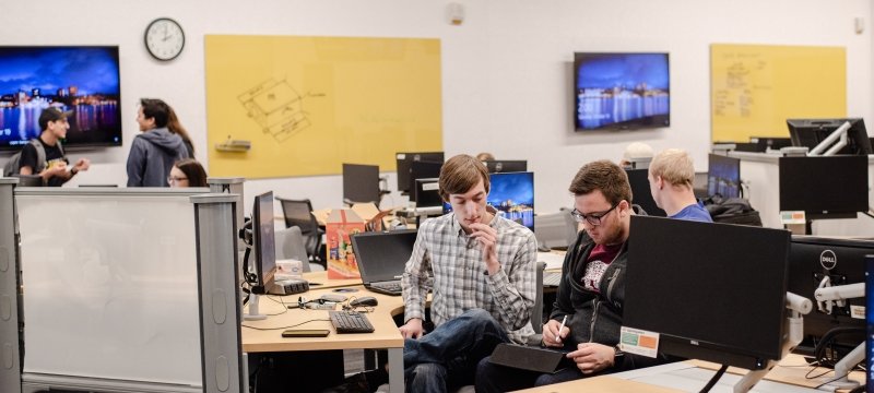 A group of students with yellow white boards and monitors and a clock reading 2 p.m. sits and stands in a learning center lab, two young men close to the camera and the others in the background.