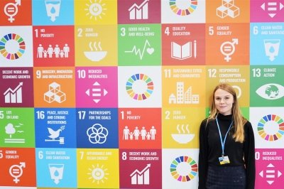 A woman stands in front of a wall of pictures depicting the United Nations Sustainable Development Goals.