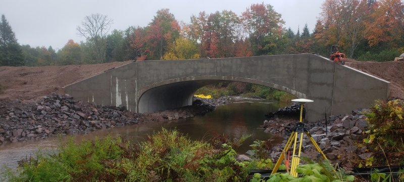 A bridge crosses a river. The ground around it is bare because the bridge was just installed. Surveying equipment is in the foreground of the photo on shore.