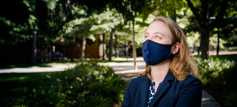A young woman with a blue face mask looks up with a forested campus and sidewalks with a brick building behind her.