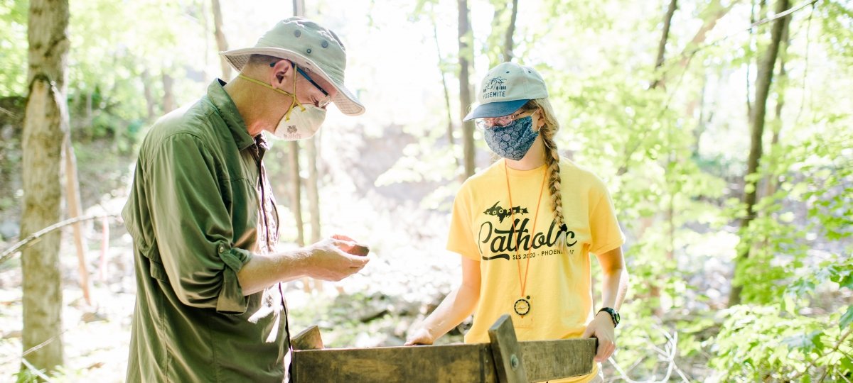 Two people wearing face coverings use a sluice box to sort through rocks and dirt. 