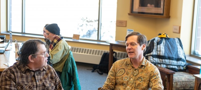 two men facing the camera and a young woman with an older man in the background talking in a library during a computer literacy session